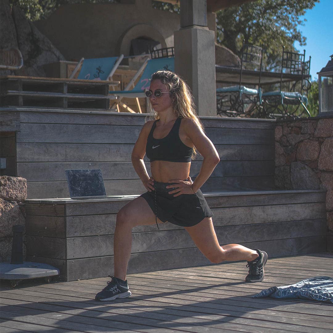 A beautiful cute looking girl wearing black sports bra and black sunglasses with black shorts doing lunges under shallow sunlight